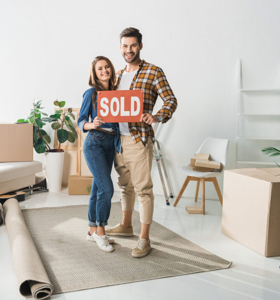 smiling couple holding sold red card at home with cardboard boxes