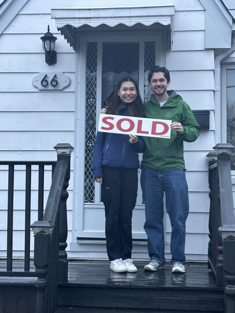 Young couple with sold sign in front of their home purchased with Nicole McKenzie, Taking you Home